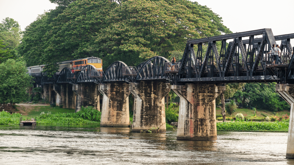 Kanchanaburi és a Kwai-folyó  fekete hídja, amint átmegy rajta egy sárga vonat